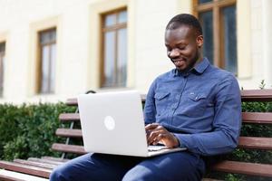 glücklicher Afroamerikanermann arbeitet an seinem Laptop, der draußen auf der Bank sitzt foto