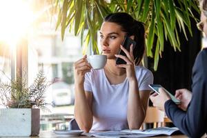 Junge charmante Frau, die mit dem Handy anruft, während sie in der Freizeit im Café sitzt, attraktive Frau mit süßem Lächeln, die sich mit dem Handy unterhält, während sie sich im Café ausruht foto