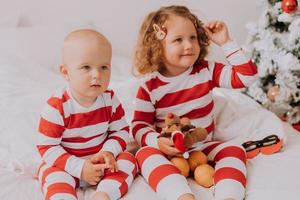 kinder in rot-weißen pyjamas probieren lustige brillen an, während der weihnachtsmann im bett sitzt. Lebensstil. bruder und schwester feiern weihnachten. Junge und Mädchen spielen zu Hause. hochwertiges Foto