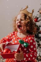 kleines lockiges mädchen in einem karnevalskleid versteckte ihr gesicht hinter glänzenden weihnachtsbaumspielzeugsternen. kind in einem roten kleid mit einem weihnachtsmanndruck auf dem hintergrund eines weihnachtsbaums. hochwertiges Foto