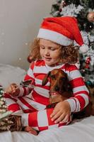 hund und kleines mädchen in rot-weißen pyjamas essen einen leckeren hausgemachten weihnachtslutscher, der im bett sitzt. Kind und ein Haustier. Baby und Dackel haben Spaß und feiern das neue Jahr. Lebensstil foto