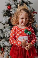 kleines lockiges mädchen in einem karnevalskleid versteckte ihr gesicht hinter glänzenden weihnachtsbaumspielzeugsternen. kind in einem roten kleid mit einem weihnachtsmanndruck auf dem hintergrund eines weihnachtsbaums. hochwertiges Foto