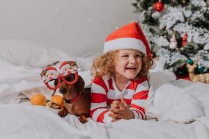 Kleines Mädchen in gestreiftem Pyjama und Weihnachtsmütze und Hund in lustigen Gläsern mit Weihnachtsmann liegen im Bett auf einem weißen Laken vor dem Hintergrund des Weihnachtsbaums. Platz für Text. hochwertiges Foto