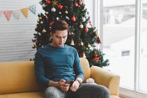 Ein einzelner Mann scheint einsam zu sein und sein Telefon in seinem mit Weihnachtsbäumen geschmückten Wohnzimmer zu benutzen foto