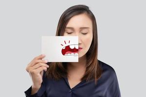asiatische frau, die ein papier mit dem gebrochenen zahnkarikaturbild seines mundes gegen den grauen hintergrund, verfallenen zahn, das konzept mit gesundheitszahnfleisch und zähnen hält foto