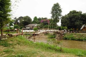 Blick auf eine Bambusbrücke über den kleinen Bergfluss mit Häusern am Ufer. pai, provinz mae hong bald, thailand. foto