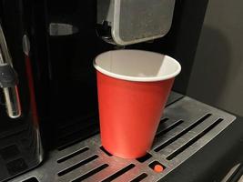 rote kaffeetasse aus pappkarton, in die morgens im büro köstlicher belebend heißer schwarzer americano-kaffee aus der kaffeemaschine gegossen wird foto