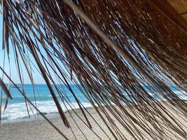 Blick auf das blaue Meer mit Salzwasser durch das gelbe trockene Stroh des Sonnenschirms am Strand im warmen östlichen tropischen Land Southern Paradise Resort. der Hintergrund foto