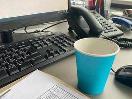 Pappblaue Kaffeetasse aus einem Café auf einem Desktop mit einem Computer mit Tastatur und Maus und Büromaterial eines Büroangestellten während des Mittagessens. geschäftliche Kaffeepause foto