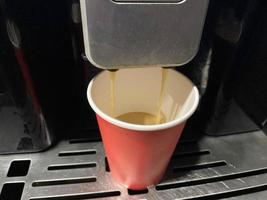 rote kaffeetasse aus pappkarton, in die morgens im büro köstlicher belebend heißer schwarzer americano-kaffee aus der kaffeemaschine gegossen wird foto