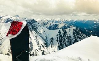 schwarzes snowboard mit weihnachtsmütze stehen im schnee schöne weiße kaukasusberge im gudauri-skigebiet top aussichtspunkt im winter. georgia skiurlaub winterurlaubsziel foto