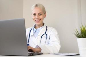 Moderne Technologie für das Gesundheitswesen. lächelnde ärztin sitzt am schreibtisch, benutzt laptop, uhr, professionelles schulungs-webinar, online, elektronisches medizinisches dokument verwalten. foto