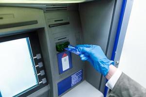 frau mit maske im gesicht und schutzhandschuhen, die ihre kreditkarte in den geldautomaten einführen, um geld zu sammeln. frau mit schutzmaske mit geldautomat foto
