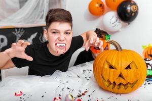 Gruseliger Junge mit Reißzähnen, der auf der Halloween-Party in die Kamera schaut. Jack o' Lantern Halloween-Kürbis auf dem Tisch foto