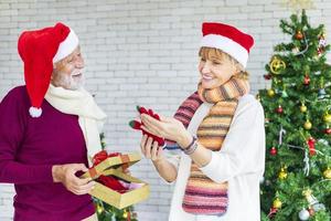 glücklich lächelndes älteres kaukasisches paar, das zusammen weihnachten feiert, während es sein geschenk in glück und aufregung zu hause mit roter weihnachtsmütze und weihnachtsbaum austauscht foto