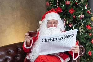 der weihnachtsmann liest weihnachtsnachrichten aus der zeitung, während er auf der sofacouch neben dem weihnachtsbaum sitzt, um lange ferien, wirtschaftliche situation und unfallverkehr, transportbericht zu erhalten