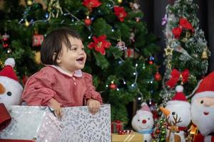 kaukasisches kleinkindbaby lächelt glücklich, während es spaß hat, um geschenkbox von den eltern zu spielen, während es in weihnachtskleid mit weihnachtsbaum auf dem rücken für das saisonfeierkonzept kleidet foto
