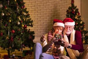 älteres kaukasisches paar, das weihnachten zusammen in glück und aufregung zu hause mit roter weihnachtsmütze und weihnachtsbaum feiert foto