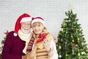 glücklich lächelndes älteres kaukasisches paar, das zusammen weihnachten feiert, während es sein geschenk in glück und aufregung zu hause mit weihnachtsmütze und weihnachtsbaum zeigt