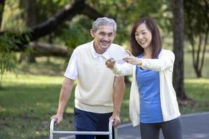 asiatischer älterer vater mit gehhilfe und tochter, die zusammen spazieren gehen, während sie im sommer selfie im park machen, um leichte übungen und physiotherapie zu nutzen foto