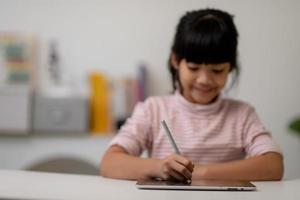 Asiatisches kleines süßes Mädchen, das den digitalen Tablet-Bildschirm auf dem Tisch berührt foto