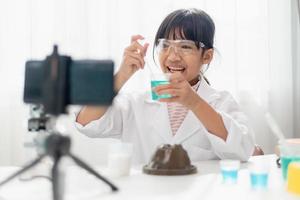 asiatisches schulmädchen, das einfache chemieexperimente macht und ein video für seine anhänger aufnimmt, junges bloggerkind, das vor der kamera für vlog posiert, kinder machen vlog für social-media-kanalkonzept. foto