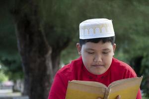 junger asiatischer muslimischer junge trägt hut, sitzt im schulpark und liest sein buch in seiner freizeit, bevor er nach hause geht, weicher und selektiver fokus. foto