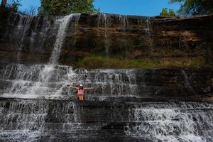 junge asiatische frau, die mit erhobenen händen sitzt und sich am wasserfall am thit mi-wasserfall, khon kaen, thailand, entspannt. foto