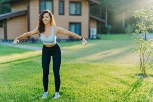 schlanke, motivierte brünette frau in kurz geschnittenem top und leggings, trainiert mit hanteln, posiert auf grünem rasen in der nähe eines privathauses, hat perfekte körperform. gesundes lebensstil- und sportkonzept foto