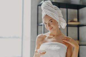 Foto einer zufriedenen jungen Frau mit gesunder, reiner Haut, trägt Körpercreme auf, spendet Feuchtigkeit mit Hautbehandlungen, steht in ein Handtuch gewickelt, lächelt sanft. persönliches pflege-, schönheits- und wellnesskonzept