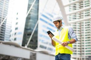 Kaukasischer Ingenieur verwendet ein Smartphone zum Reden, trägt eine gelbe Weste und einen großen Schutzhelm und hält andererseits den weißen Grundriss in der Baustelle der Innenstadt. foto