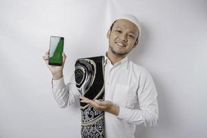 ein Porträt eines glücklichen asiatischen muslimischen Mannes, der lächelt, während er Kopienraum auf seinem Telefon zeigt, isoliert durch weißen Hintergrund foto