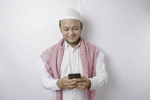 ein Porträt eines glücklichen asiatischen muslimischen Mannes, der lächelt, während er sein Telefon hält, isoliert durch weißen Hintergrund foto