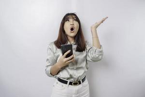 schockierte asiatische Frau mit salbeigrünem Hemd, die auf den Kopierbereich auf ihr zeigt, während sie ihr Telefon hält, isoliert durch weißen Hintergrund foto