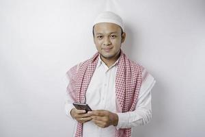 ein Porträt eines glücklichen asiatischen muslimischen Mannes, der lächelt, während er sein Telefon hält, isoliert durch weißen Hintergrund foto