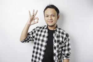 Aufgeregter asiatischer Mann mit Tartan-Hemd, der eine ok-Handgeste gibt, die durch einen weißen Hintergrund isoliert ist foto