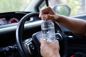 asiatische fahrerin hält kaltes wasser zum trinken im auto, gefährlich und riskiert einen unfall.