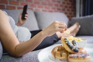 fette frau, die auf dem sofa liegt, smartphone hält und kartoffelchips isst, überessendes konzept foto