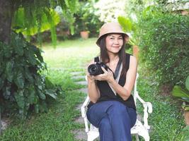 asiatische frau, mit hut, schwarzem oberteil ohne ärmel und blauer jeanshose, sitzt auf einem weißen stuhl im garten und hält eine dslr-kamera und lächelt glücklich. foto