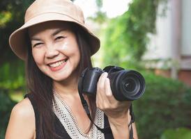 asiatische frau mit hut und schwarzem oberteil ohne ärmel, die im garten steht und eine dslr-kamera hält und glücklich lächelt. foto