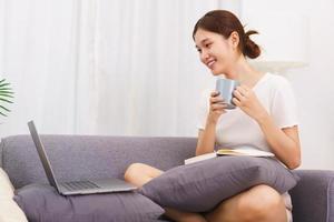 Lebensstil im Wohnzimmerkonzept, asiatische Frau sitzt auf der Couch, um Kaffee zu trinken und Filme anzusehen