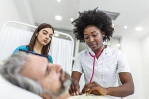 Kranker Patient mit afroamerikanischer Ärztin hört seine Brust mit Stethoskop in der Notaufnahme des Krankenhauses. Arzt und Krankenschwester kommen, um ältere Patienten im Krankenzimmer zu besuchen. foto