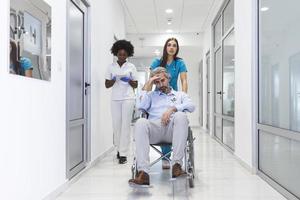 Krankenschwester mit Scrubs, die Patienten im Rollstuhl durch die Lobby eines modernen Krankenhausgebäudes rollt. ärzte und krankenschwester im krankenhauskorridor mit älterem männlichen patienten im rollstuhl