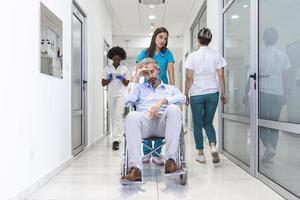 Krankenschwester mit Scrubs, die Patienten im Rollstuhl durch die Lobby eines modernen Krankenhausgebäudes rollt. ärzte und krankenschwester im krankenhauskorridor mit älterem männlichen patienten im rollstuhl