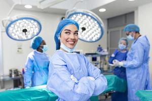 Nahaufnahme einer Chirurgin, die mit Kollegen, die im Hintergrund im Operationssaal auftreten, in die Kamera blickt. das Konzept der Medizin foto