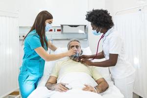 junge ärztin und krankenschwester mit chirurgischer maske, die einen älteren männlichen patienten mit einer überdruck-sauerstoffmaske überprüft, um das atmen in einem krankenhausbett während der covid-19-pandemie zu unterstützen foto