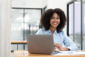 lächelnde junge afrikanische Unternehmerin, die an einem Schreibtisch in ihrem Heimbüro sitzt und online mit einem Laptop arbeitet foto