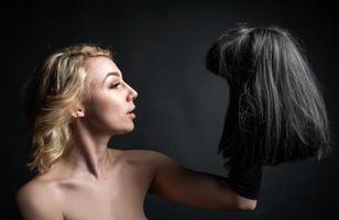 eine junge blonde Frau mit Perücke, die eine schwarze Perücke in der Hand hält. auf schwarzem Hintergrund foto