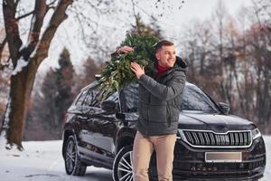 Vorderansicht. mann mit kleiner grüner tanne ist draußen in der nähe seines autos. Konzeption von Feiertagen foto