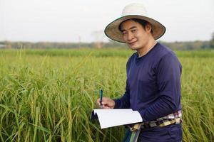 gutaussehender asiatischer Bauer ist auf Reisfeld, trägt Hut, blaues Hemd, hält Notizbuchpapier, inspiziert Wachstum und Krankheit von Pflanzen. Konzept, landwirtschaftliche Forschung und Studie zur Entwicklung von Nutzpflanzen. foto